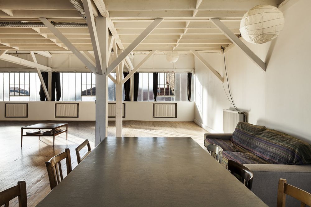 A long open loft with white beams, industrial windows, a long table and a sagging sofa